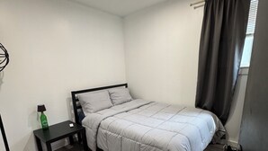 A bedroom with a bed, a nightstand, a lamp, a basket, and a window with curtains. - Modern Stay By the Falls – Renovated & Walkable! (Niagara Falls)