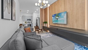 Ein modernes Wohnzimmer mit einem grauen Ecksofa, einer Holzwand, einem Wand-Fernseher und einer Deckenleuchte.