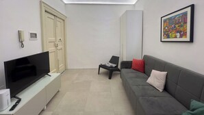 A modern living room with a flat-screen TV on a white cabinet, a grey sofa with red and white cushions, a small table with a laptop, and a colorful painting on the wall. - [ Martiry Suite Apartment ] + Self Check-in Late (Catania)