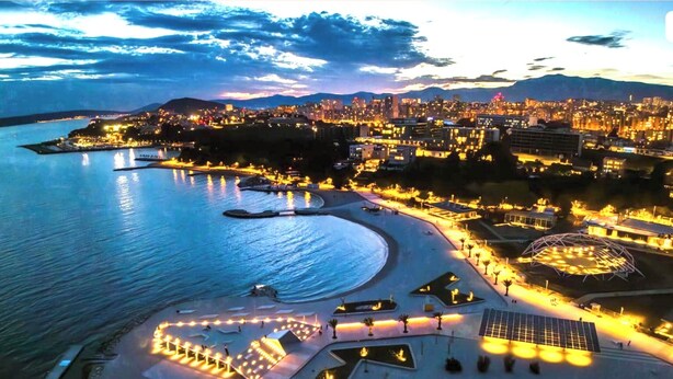 An aerial view of a coastal cityscape with a well-lit beachfront promenade, a large circular structure, and a distant mountain range. - Stella's beachfront aparthotel (Split)