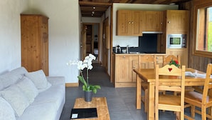 A cozy living room with a sofa, a coffee table, and a potted orchid. A wooden dining table with chairs is in the background, and a kitchen area with wooden cabinets and a microwave is visible. - Entire chalet with indoor pool jacuzzi sauna steam room (Taninges)