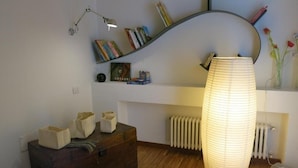A room with a wooden floor, a dark wooden dresser, a white radiator, a large white vase lamp, and a curved bookshelf with books and a picture frame. - Puccini- un bel dì (Cagliari)