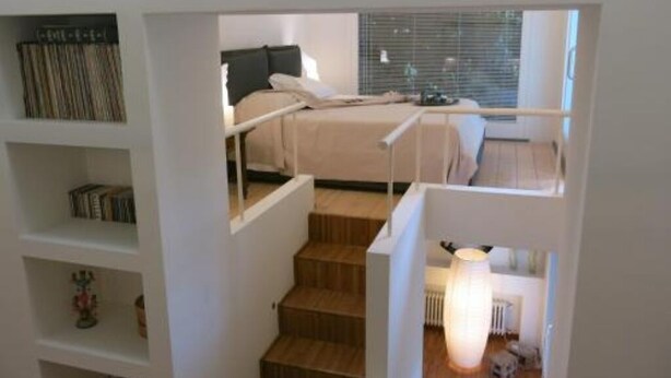 A bedroom with a bed, a nightstand, a lamp, and a window with blinds. - Puccini- un bel dì (Cagliari)