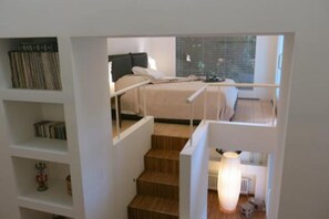 A bedroom with a bed, a nightstand, a lamp, and a window with blinds. - Puccini- un bel dì (Cagliari)