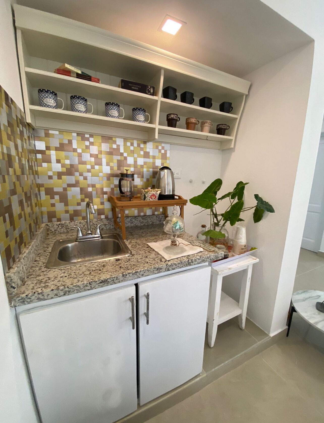 A compact kitchen with a granite countertop, a coffee maker, a kettle, and a small shelf with coffee mugs.