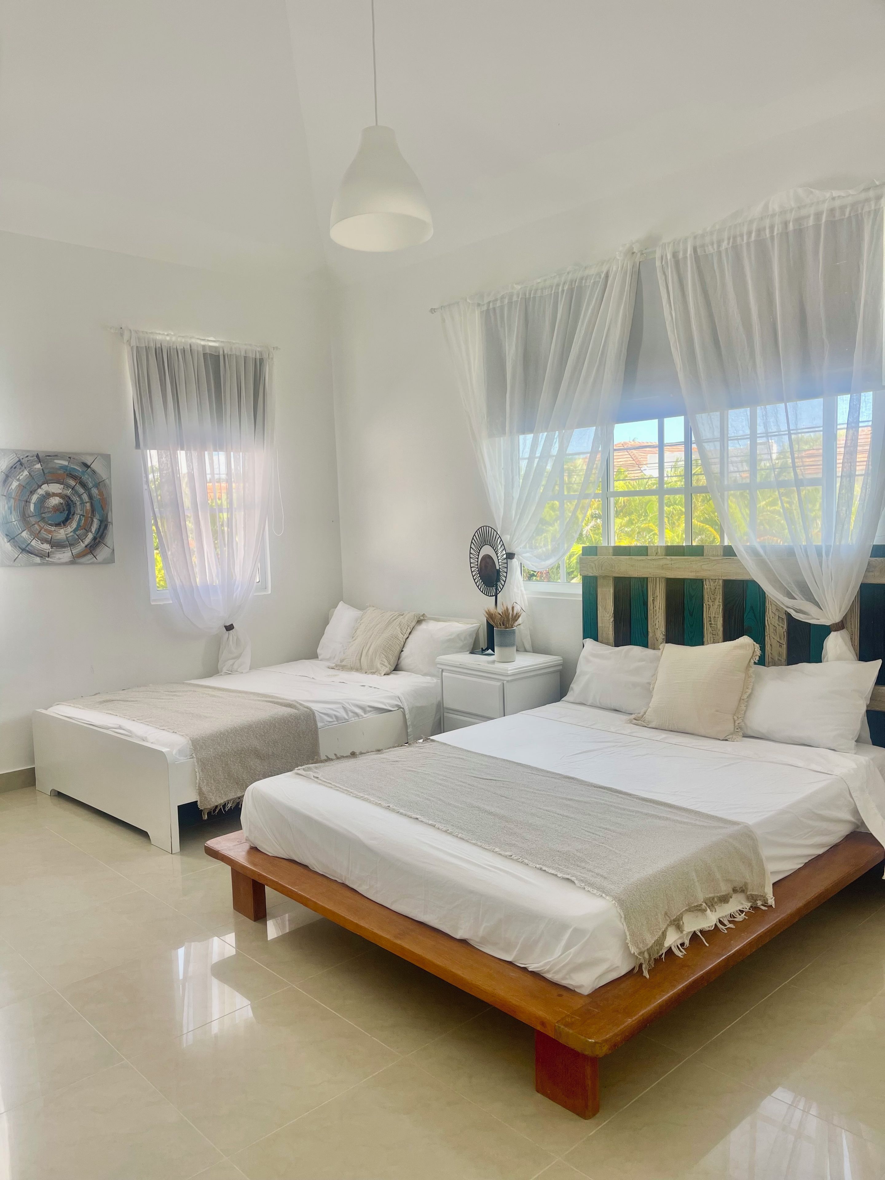 A bedroom with two single beds, a wooden bench, a bedside table with a lamp, and sheer curtains.