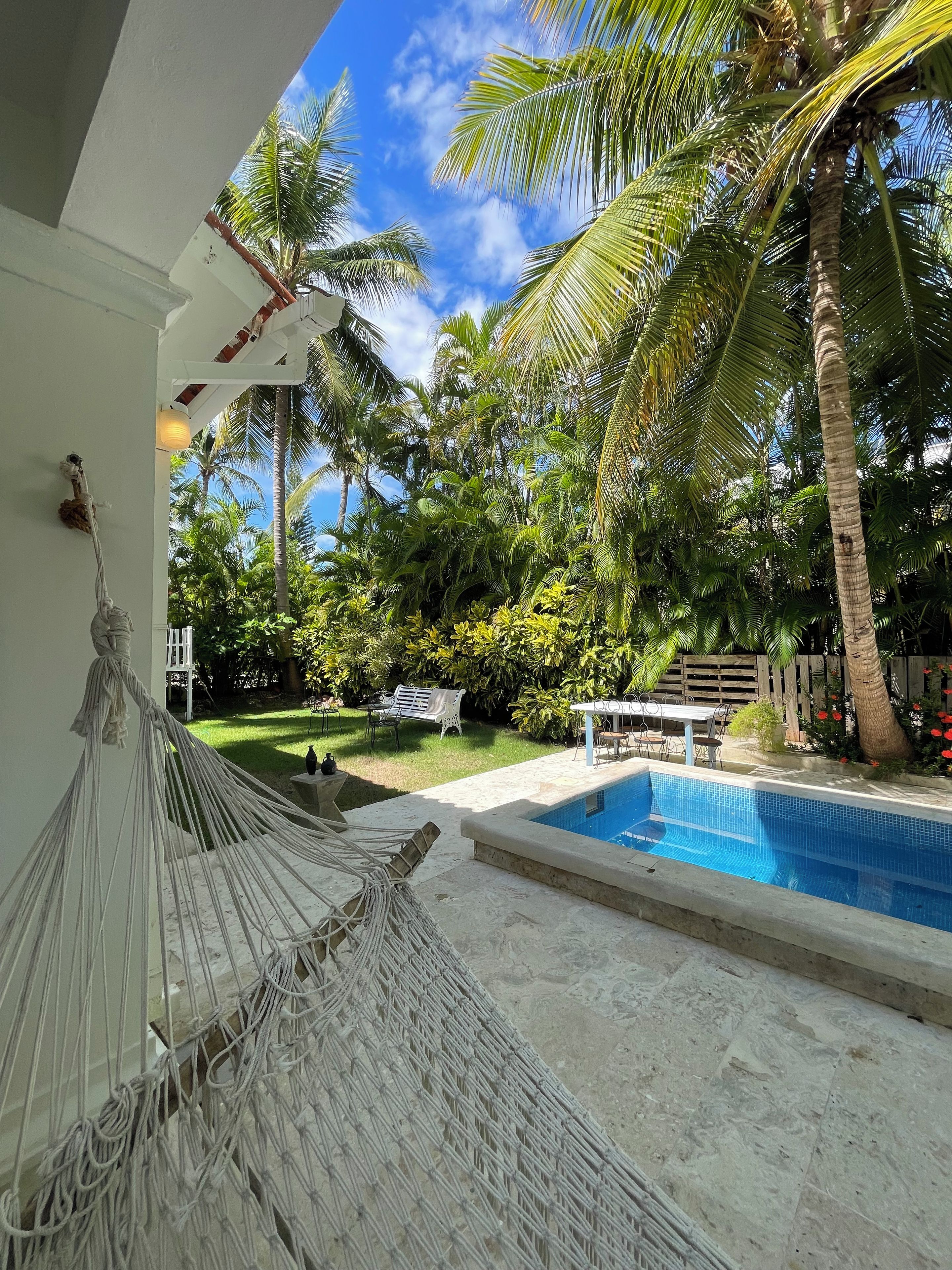 Un área de piscina con una hamaca, palmeras y una mesa con sillas.