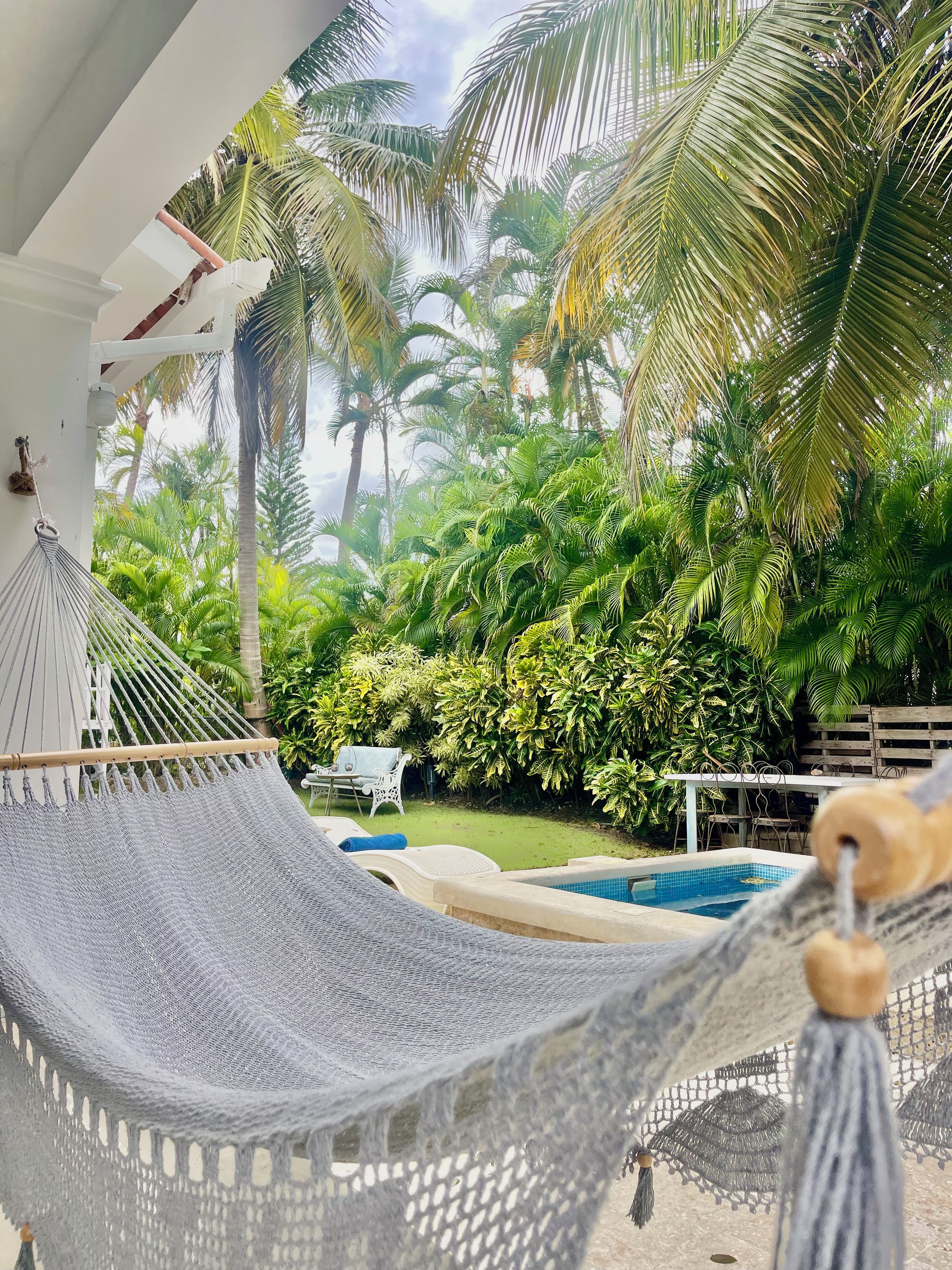A hammock under a white structure with a view of a pool and lush greenery.