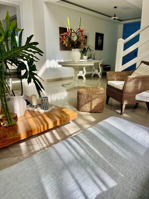 Une pièce bien éclairée, avec une table à manger, des chaises en osier, une plante en pot et un vase rempli de fleurs.