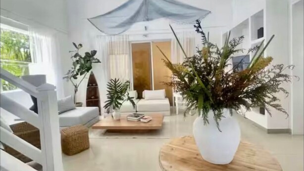 Een ruime woonkamer met een witte bank, een houten salontafel en een grote witte vaas met planten.