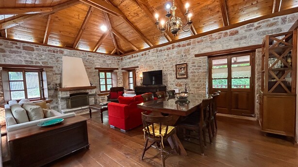 A rustic interior with a stone wall, wooden ceiling, a fireplace, a television, and a dining area. - Villa Acroria in Karpenisi (Karpenisi)