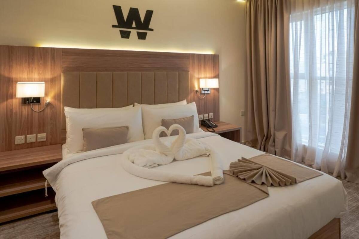 A hotel room with a large bed, a headboard with a 'W' logo, bedside lamps, and a window with curtains.