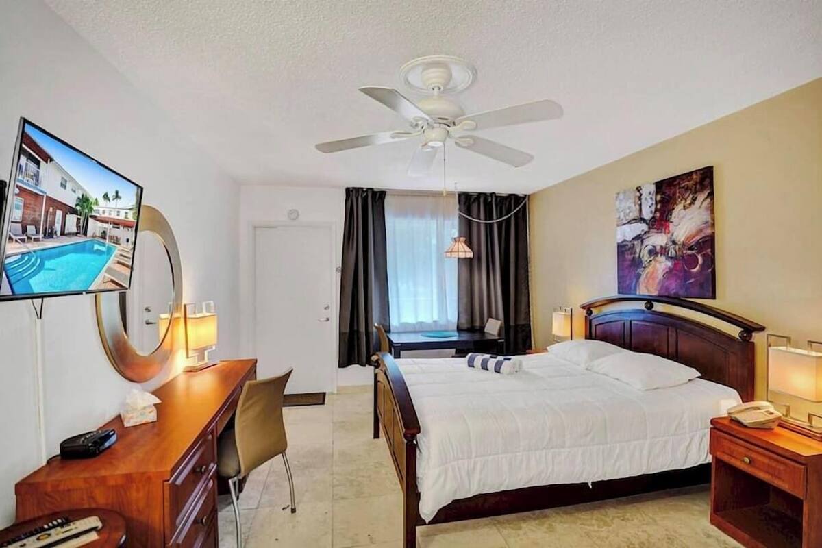 Habitación de hotel con cama, escritorio, silla, televisor, ventilador de techo y obra de arte en la pared.