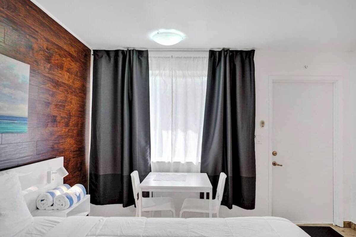 Una habitación de hotel con una cama, una mesita con sillas, un cuadro en la pared y una puerta.