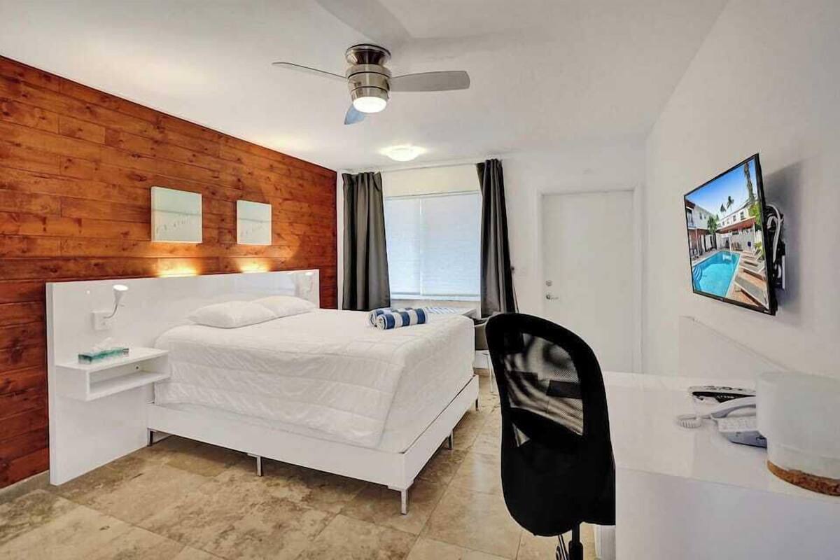 Un dormitorio con una cama, un escritorio, una silla, un ventilador de techo y un televisor en la pared.