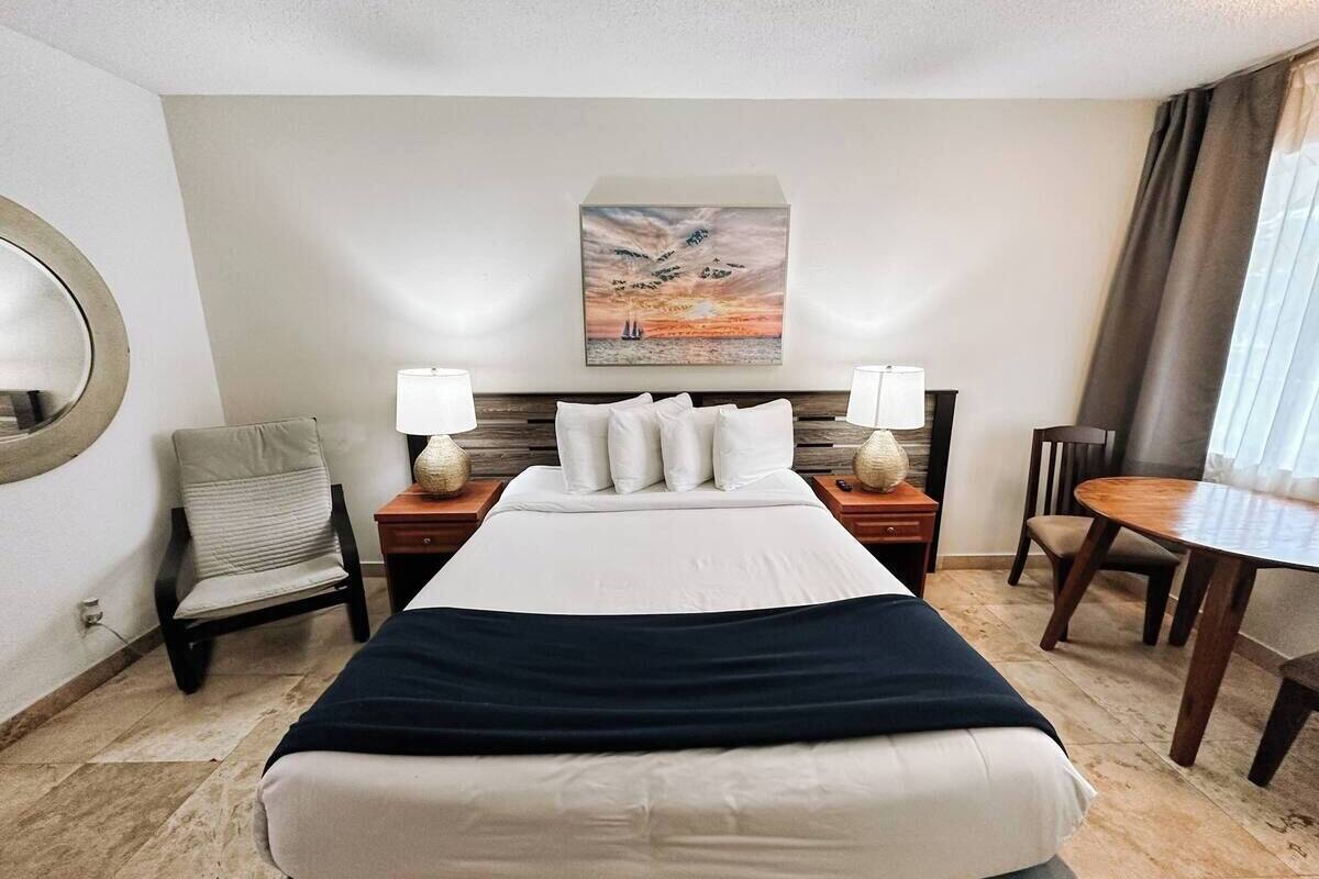 Una habitación de hotel con una cama grande, dos mesitas de noche con lámparas, una silla, una mesa redonda y un cuadro en la pared.