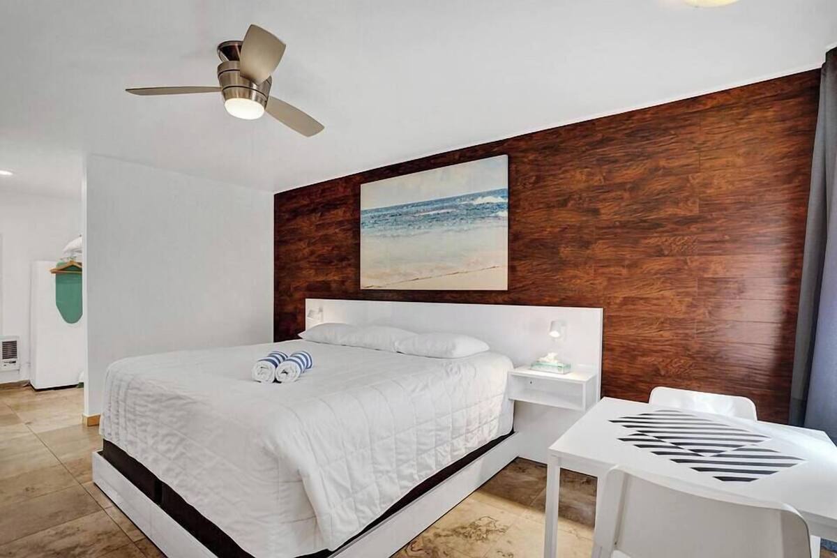 Una habitación de hotel moderna con una cama grande, un ventilador de techo, un panel decorativo de madera, un cuadro de una playa y una pequeña mesa blanca.