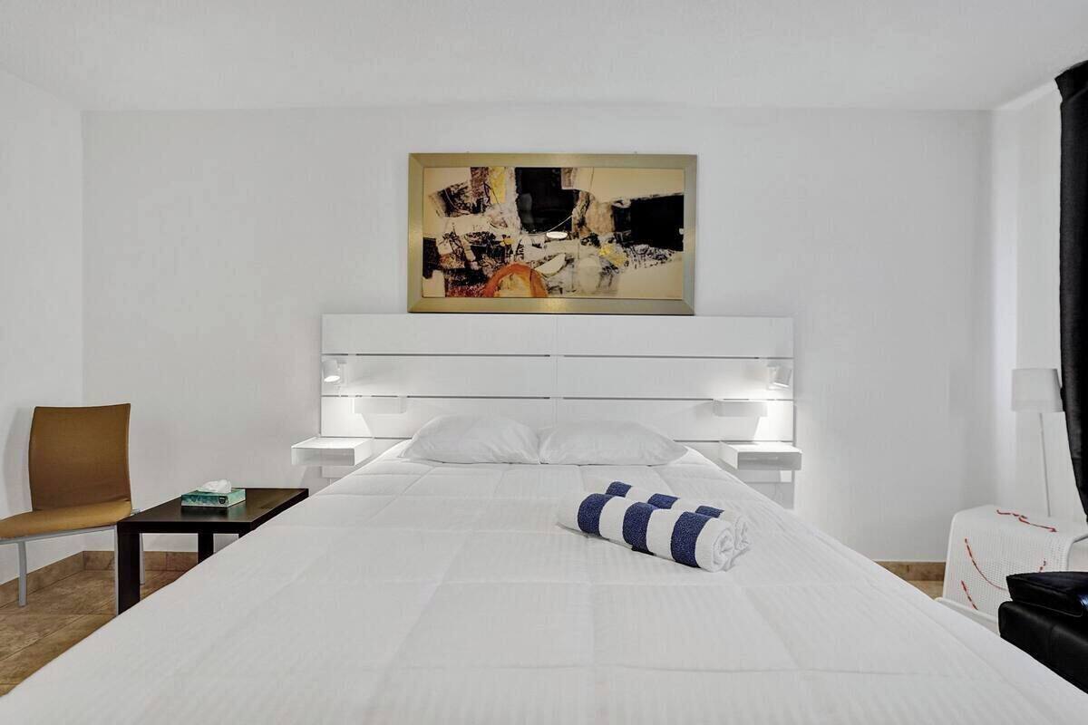 Una habitación de hotel moderna con una cama grande, cabecera blanca, una mesita, una silla y un cuadro en la pared.