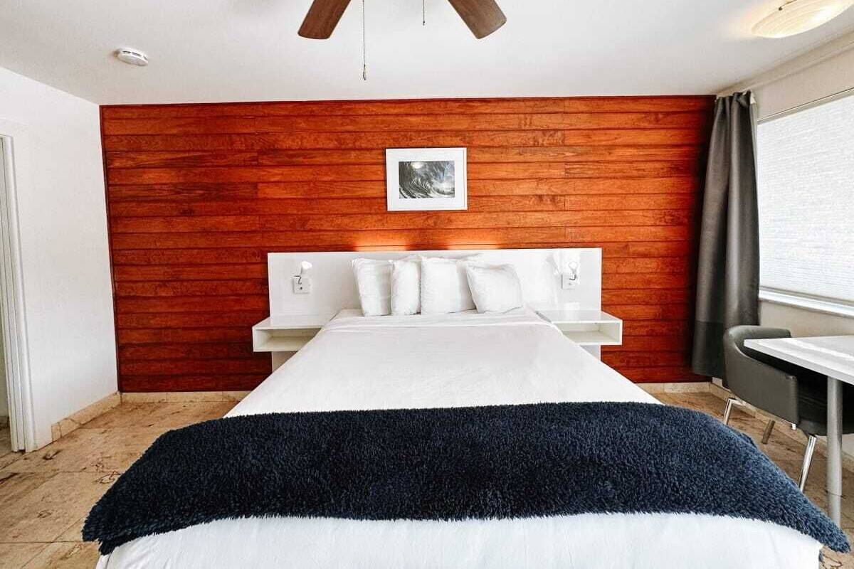 Habitación de hotel con pared de madera decorativa, cama con ropa blanca, ventilador de techo, mesita de noche y ventana con cortinas.
