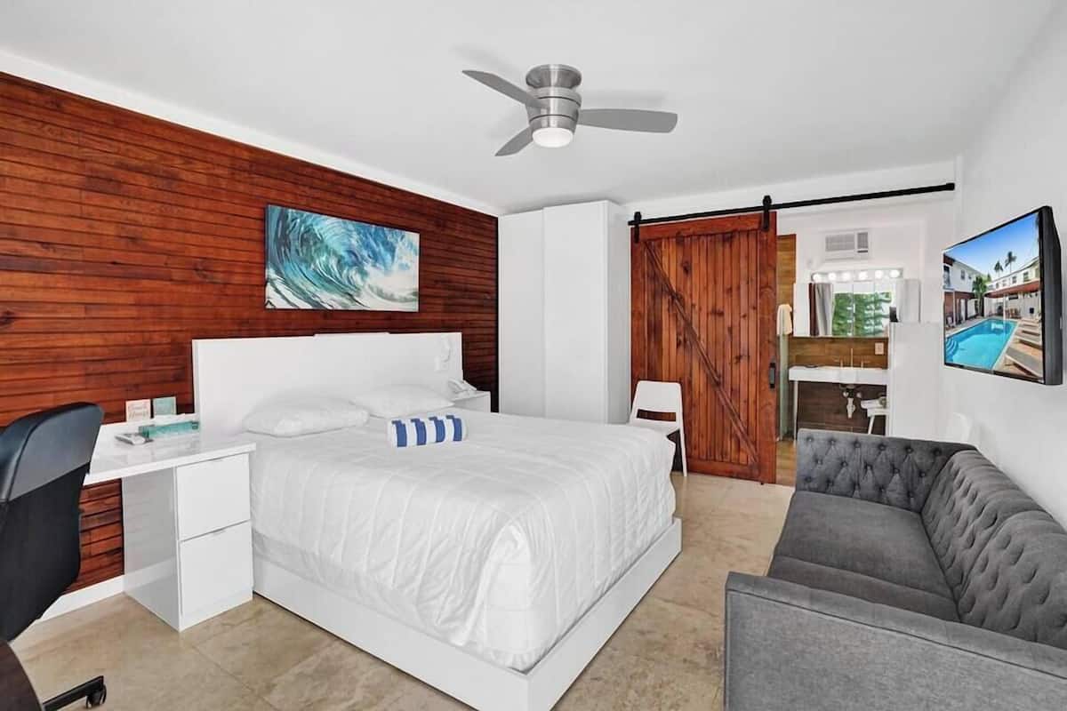 Un dormitorio con cama, escritorio, silla, sofá gris, ventilador de techo y televisor montado en la pared.
