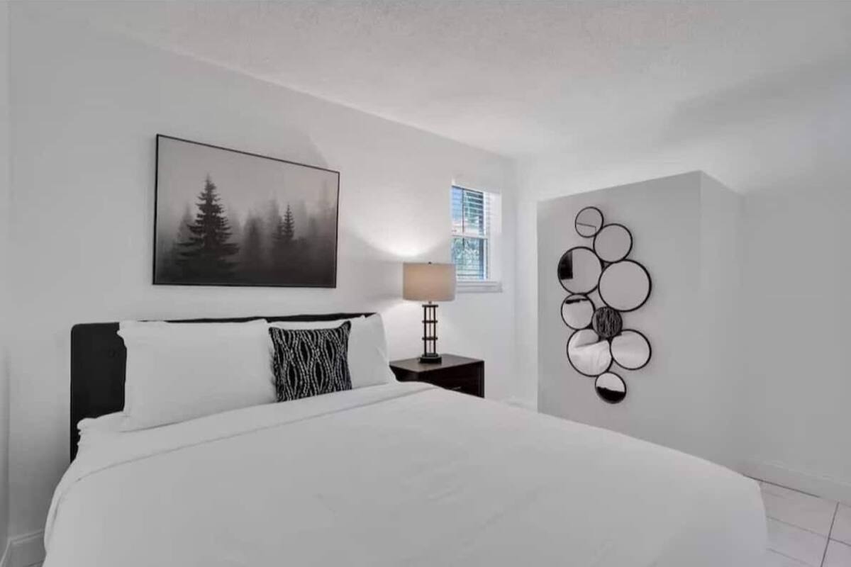 Un dormitorio con una cama grande, una mesita de noche con lámpara, una obra de arte circular colgada en la pared y un cuadro enmarcado de un paisaje forestal.