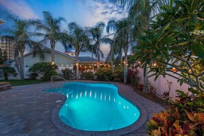 A swimming pool with a curved edge, surrounded by palm trees and a patio area with string lights. - Casa Honu (New Smyrna Beach)