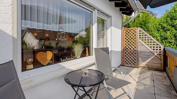 Un patio con tavolo e sedie, una recinzione in legno e una finestra con vista su una casa e vegetazione.