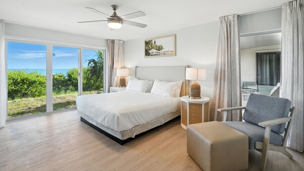 A spacious bedroom with a large bed, a ceiling fan, a view of the outdoors, and a seating area with a chair and ottoman. - Lighthouse Resort and Club (Sanibel)