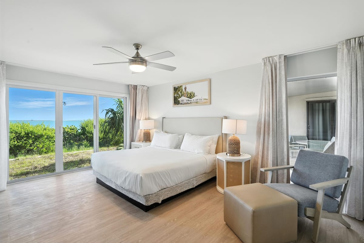 A spacious bedroom with a large bed, a ceiling fan, a view of the outdoors, and a seating area with a chair and ottoman.