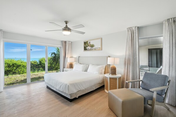 Une chambre spacieuse avec un grand lit, un ventilateur de plafond, une vue sur l’extérieur et un coin salon comprenant un fauteuil et un pouf.