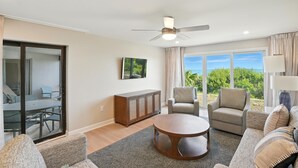 A spacious living room with a ceiling fan, a round coffee table, a sofa, a chair, and a view of the outdoors through large windows. - Lighthouse Resort and Club (Sanibel)