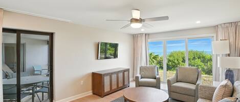 A spacious living room with a ceiling fan, a round coffee table, a sofa, a chair, and a view of the outdoors through large windows.