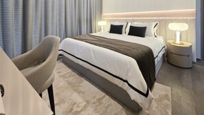 A modern hotel room with a large bed, a bedside table with a lamp, a chair, and a large window with curtains.