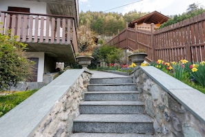 Un escalier en pierre mène à un balcon en bois d’où l’on peut admirer un jardin et un bâtiment entouré d’une clôture en bois.