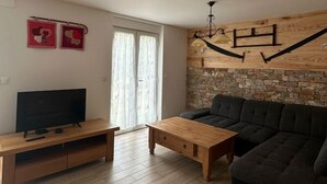 Ein Wohnzimmer mit Couchtisch aus Holz, einer dunklen Couch, einem Fernseher auf einem Holzständer und einer Steinwand mit sichtbarem Holzbalken.