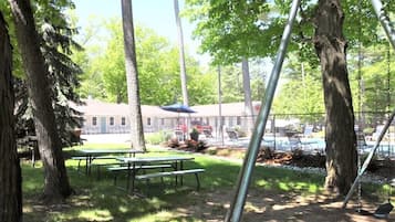 Un área de picnic con mesas y bancos, rodeada de árboles, con un edificio y una piscina al fondo.