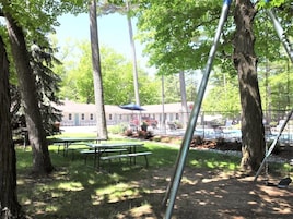 Un área de picnic con mesas y bancos, rodeada de árboles, con un edificio y una piscina al fondo.