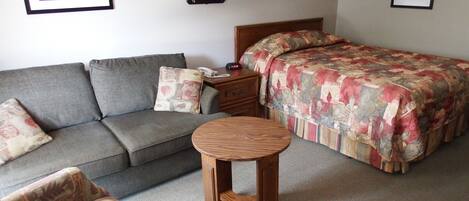 Una habitación con un sofá gris, un sillón estampado, una mesita de madera, una cama con colcha floral y dos lámparas fijadas a la pared.