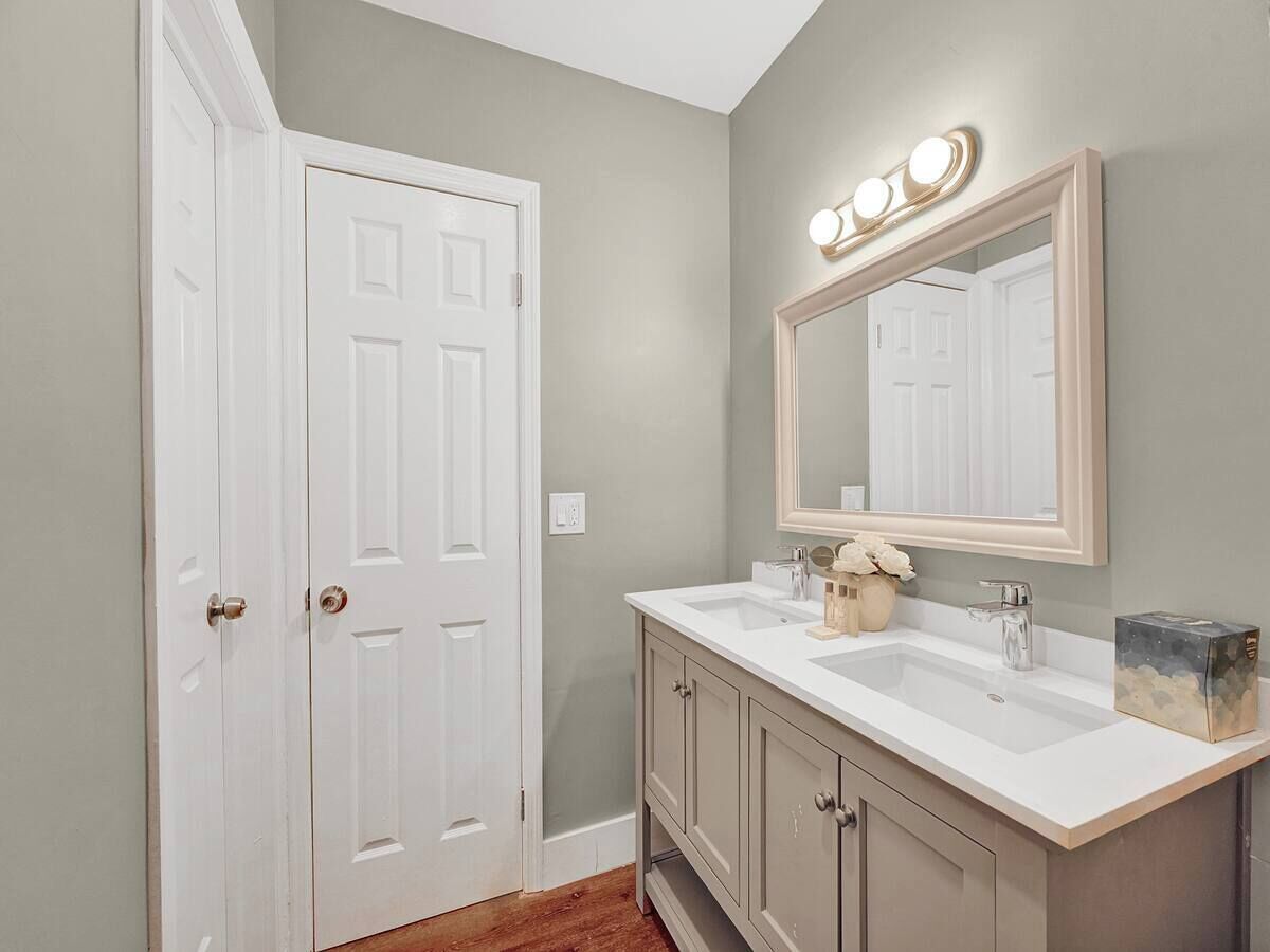 A bathroom with a double vanity, a large mirror with lights, and a white door.