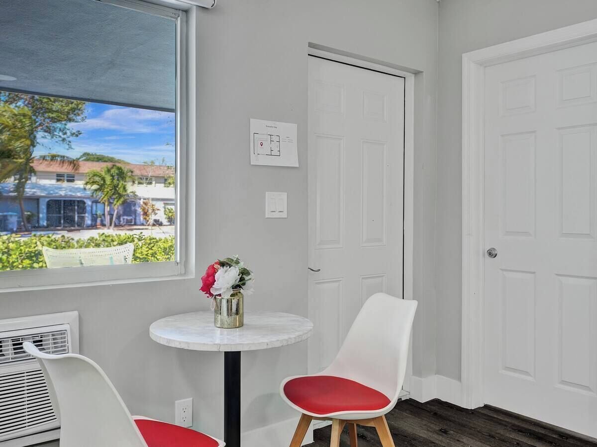 Una habitación con una puerta blanca, una silla blanca con cojín rojo, una mesita redonda con un florero y una ventana que ofrece vistas a edificios y vegetación.