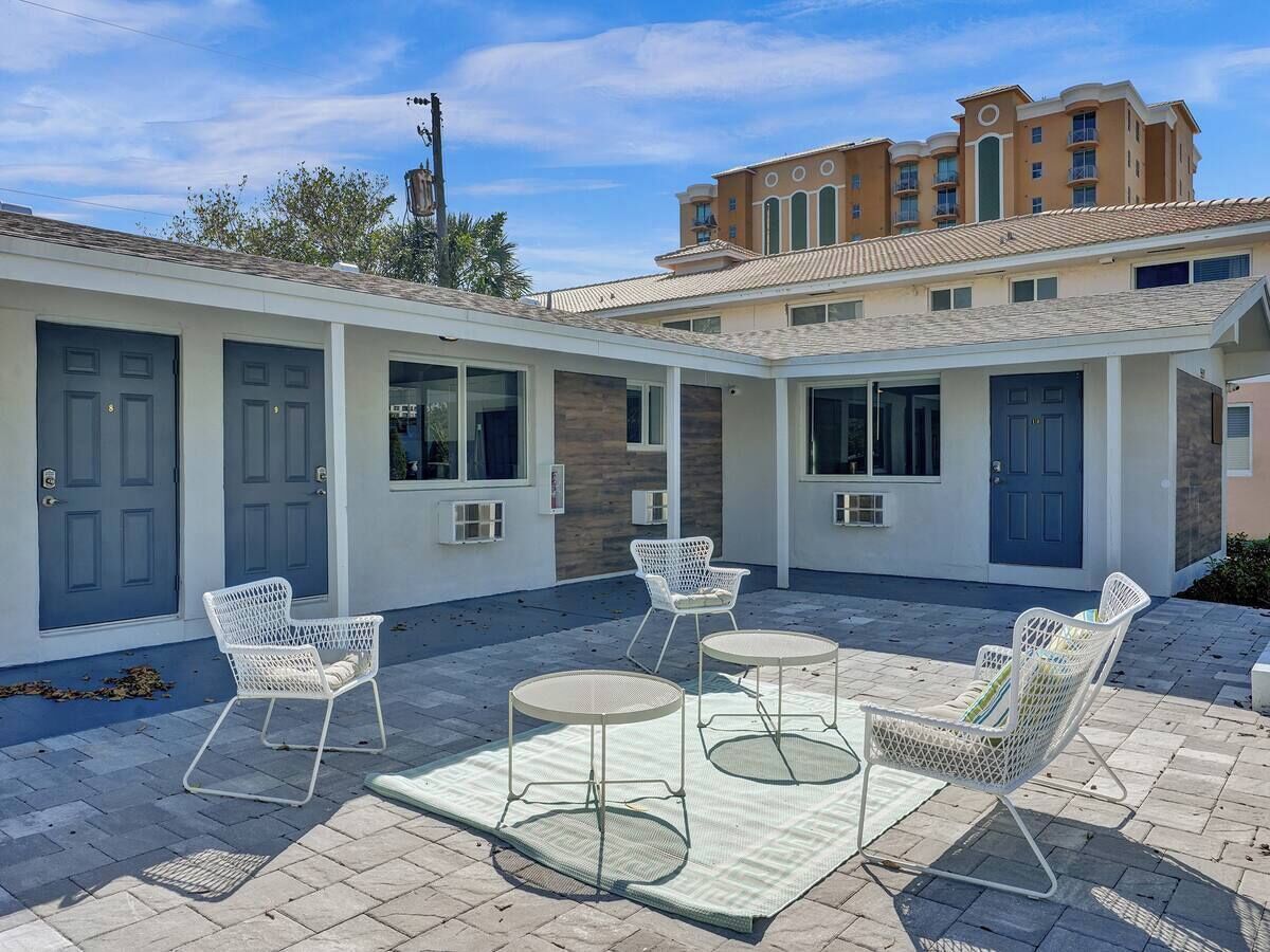 A courtyard with outdoor seating, including white chairs and round tables, and a building with multiple doors and windows in the background.