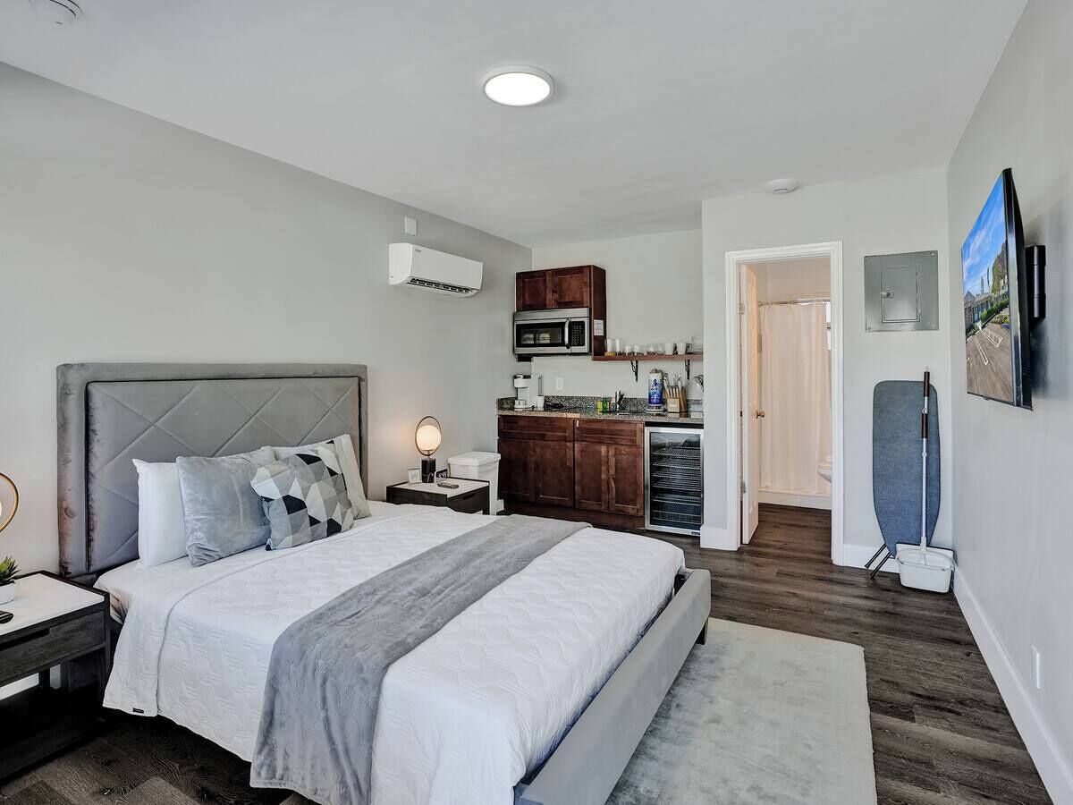 A modern bedroom with a large bed, a nightstand, a bedside table, a built-in microwave, a small kitchen area, and a door leading to another room.