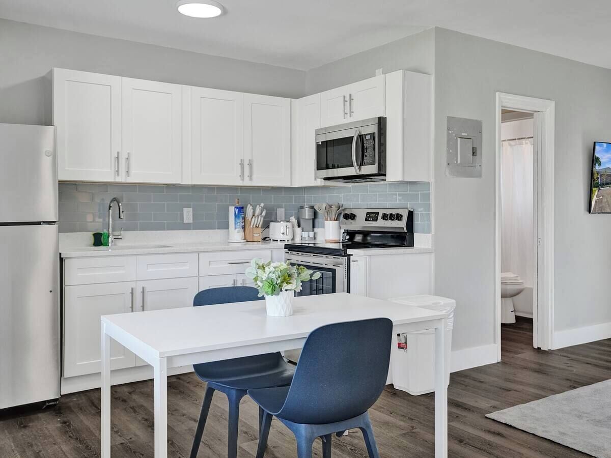 A modern kitchen with white cabinets, a stainless steel refrigerator, a microwave, and an oven. There is a dining table with two chairs and a potted plant on it.