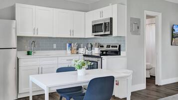 A modern kitchen with white cabinets, a stainless steel refrigerator, a microwave, and an oven. There is a dining table with two chairs and a potted plant on it.