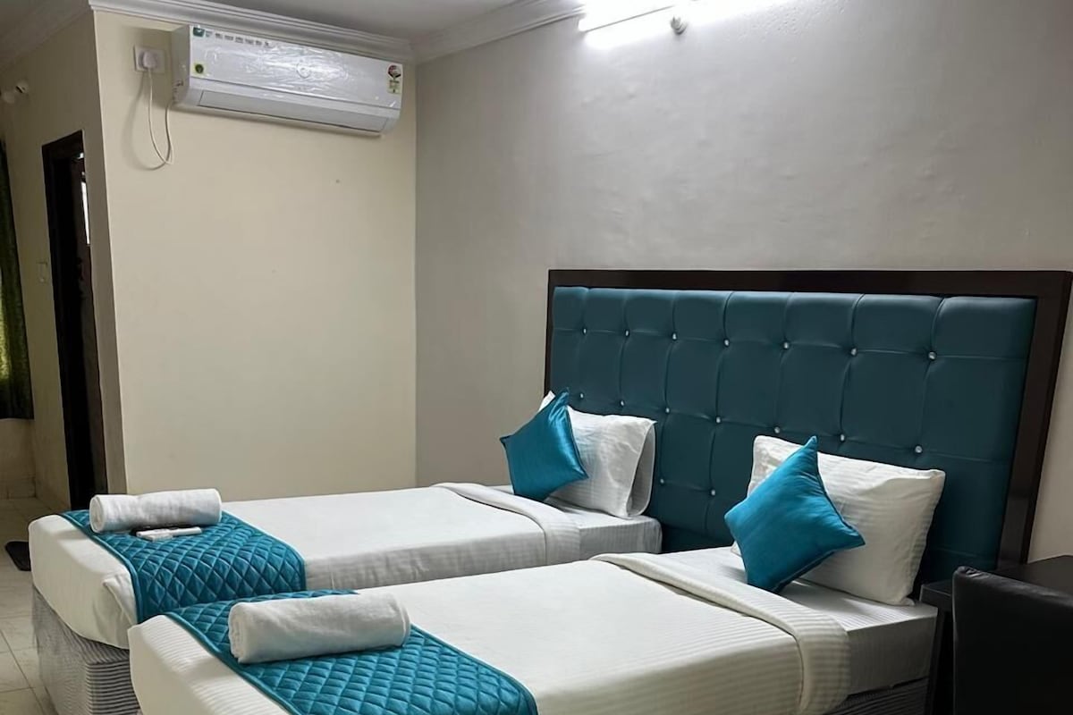 Une chambre d’hôtel avec deux lits, chacun recouvert d’une courtepointe matelassée de couleur turquoise et agrémenté de draps blancs. La chambre est équipée d’un ventilateur de plafond, d’un climatiseur et d’une petite table avec une chaise.