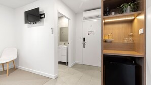 A modern hotel room with a white chair, a flat-screen TV mounted on the wall, a built-in air conditioning unit, a wooden shelf with decorative items, and a compact black refrigerator. - Modern Queen Studio - 6 Min Walk to Callala Beach (Callala Beach)