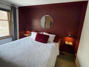 Una habitación de hotel con una cama grande, un espejo redondo en la pared, dos mesitas de noche con lámparas y una ventana con cortinas.