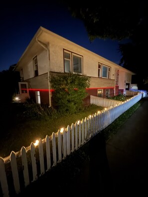 A two-story house with a white picket fence, a small garden, and a red-lit pathway leading to the front door. - Walking Distance to Downtown - 3 Comfy King Beds, Hot Tub, Sauna, Merlot House (Calgary)