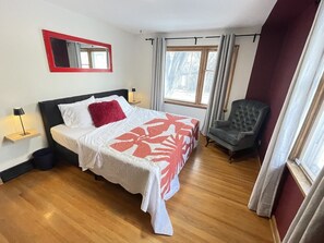 Un dormitorio con cama, una silla, una mesita de noche, un espejo y una ventana con cortinas.
