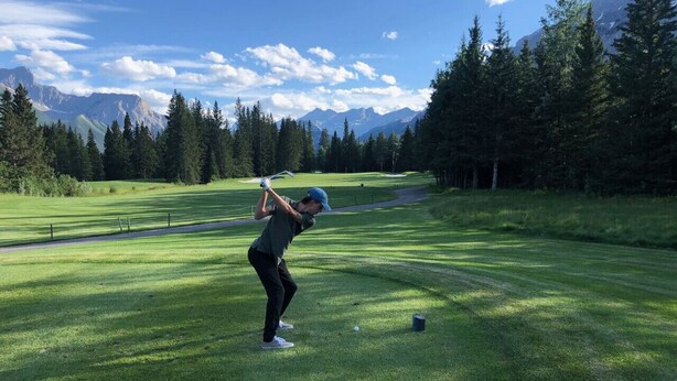 Una persona juega al golf en un campo bien cuidado, con cielo despejado y montañas al fondo.